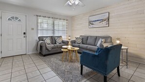A living room with a grey sofa, a blue armchair, a small round table, and a ceiling fan. - Sandy Sands Retreat! Just 1 min to the beach! (Gulfport)
