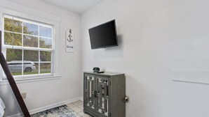 A room with a window, a flat-screen TV mounted on the wall, and a grey cabinet with glass doors. - Sandy Sands Retreat! Just 1 min to the beach! (Gulfport)