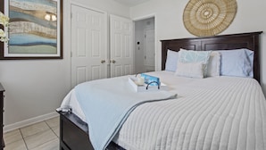 A bedroom with a large bed, a framed painting, a decorative wall hanging, and a bedside table with a lamp. - Sandy Sands Retreat! Just 1 min to the beach! (Gulfport)