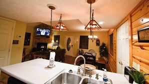 Una cocina con una isla grande, pileta de acero inoxidable e iluminación colgante. Hay un televisor montado sobre la isla y una chimenea al fondo.