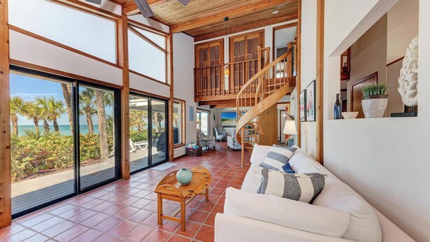 A spacious living area with a wooden staircase, large windows overlooking a beach, and a cozy seating area with a white sofa and a small wooden table.