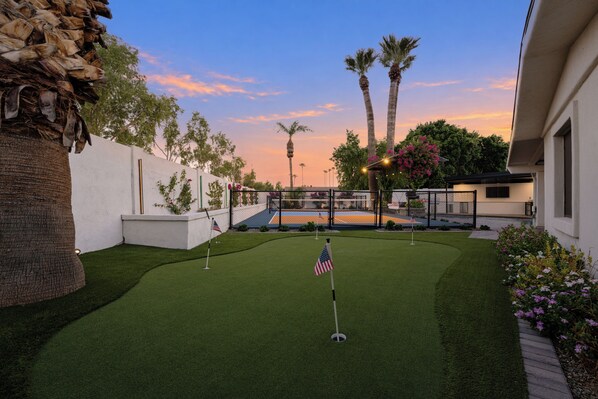 A well-maintained artificial grass golf course with a flag, palm trees, and a tennis court in the background.