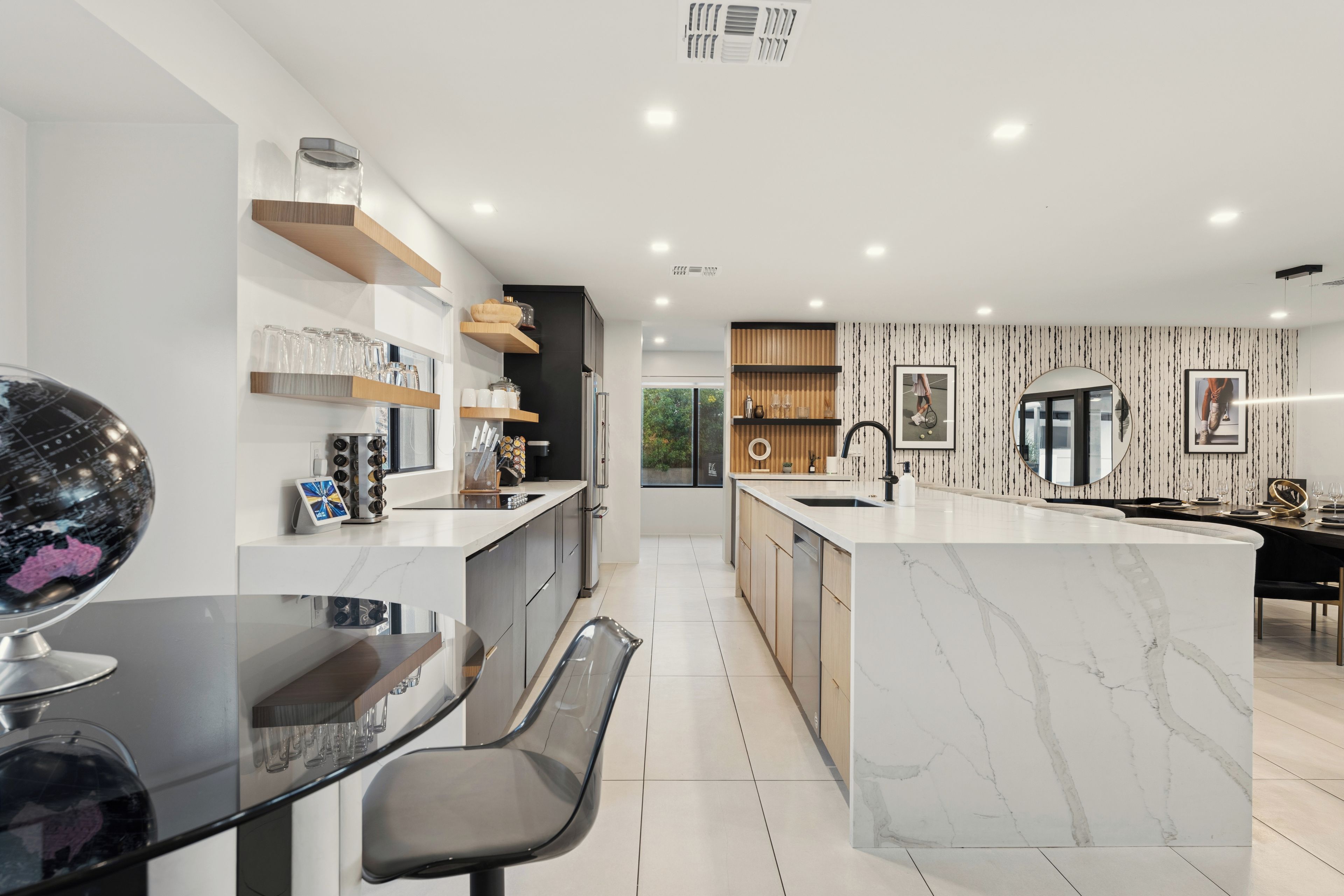 Una cocina moderna con una isla de mármol, estantes de madera y un comedor con mesa y sillas de vidrio.