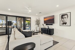 A modern living room with a sofa, a coffee table, and a TV. There is a large window with a view of the outdoors.
