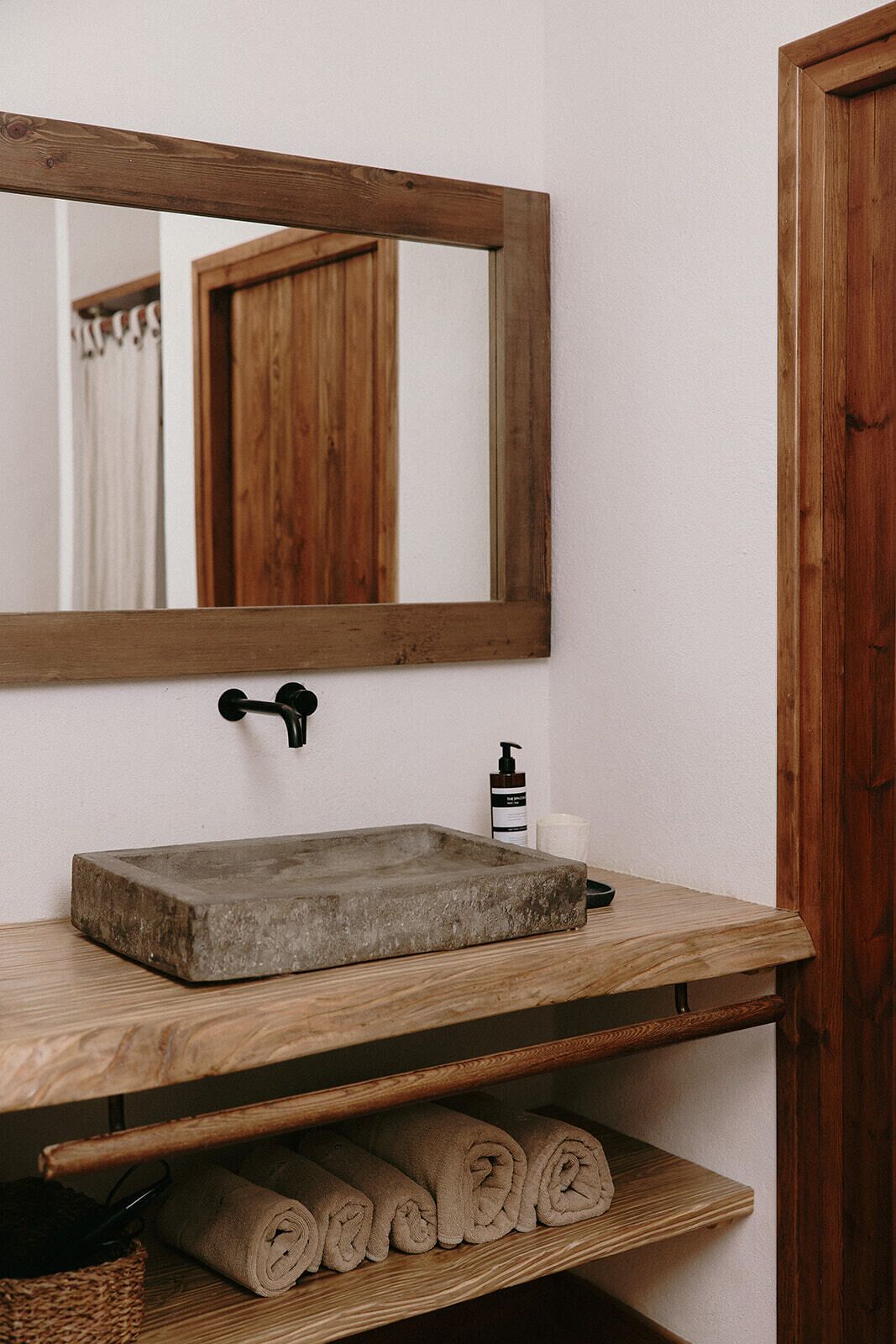 Un baño con tocador de madera, un lavamanos rectangular, un espejo, un perchero para toallas y una puerta.