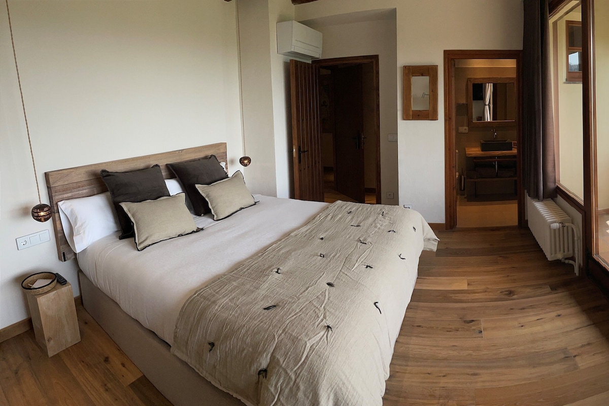 Un dormitorio con una cama grande, piso de madera, baño con lavamanos y espejo, y un balcón con vistas a las montañas.