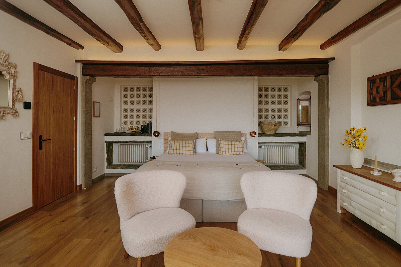 A bedroom with a double bed, two armchairs, a small round table, a dresser, and a mirror.