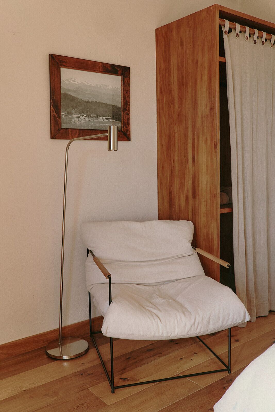 A room with a wooden wardrobe, a white chair, a floor lamp, and a framed picture on the wall.