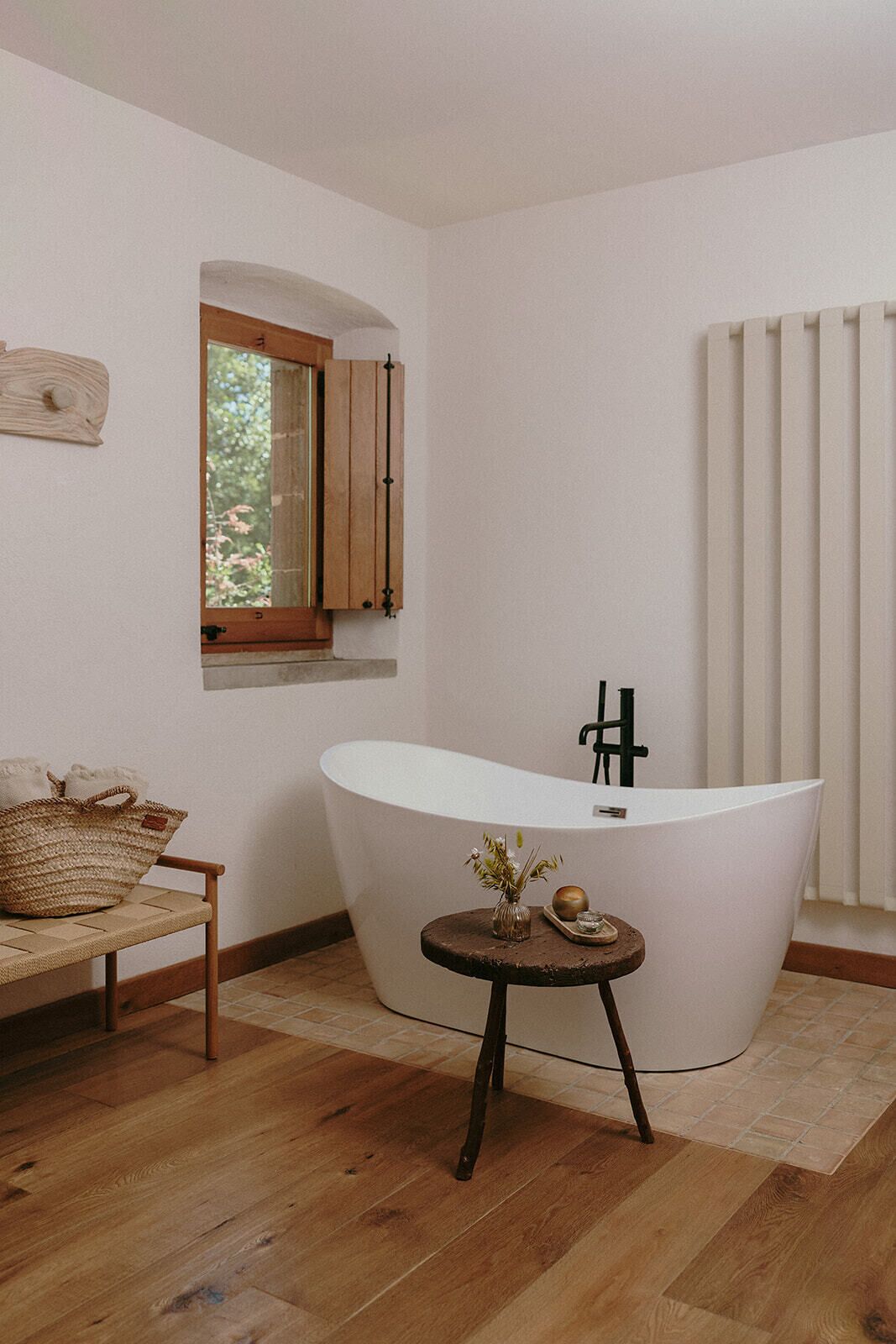 Un baño moderno con una bañera independiente, un banco de madera, una mesita redonda con un florero y una ventana con contraventanas de madera.