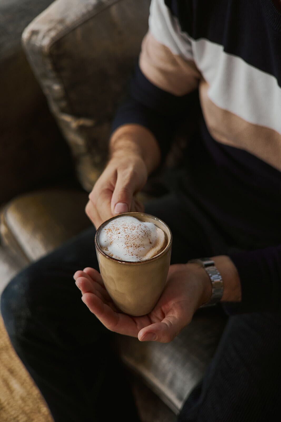 Una persona agafa una tassa de cafè amb escuma, asseguda en un sofà.