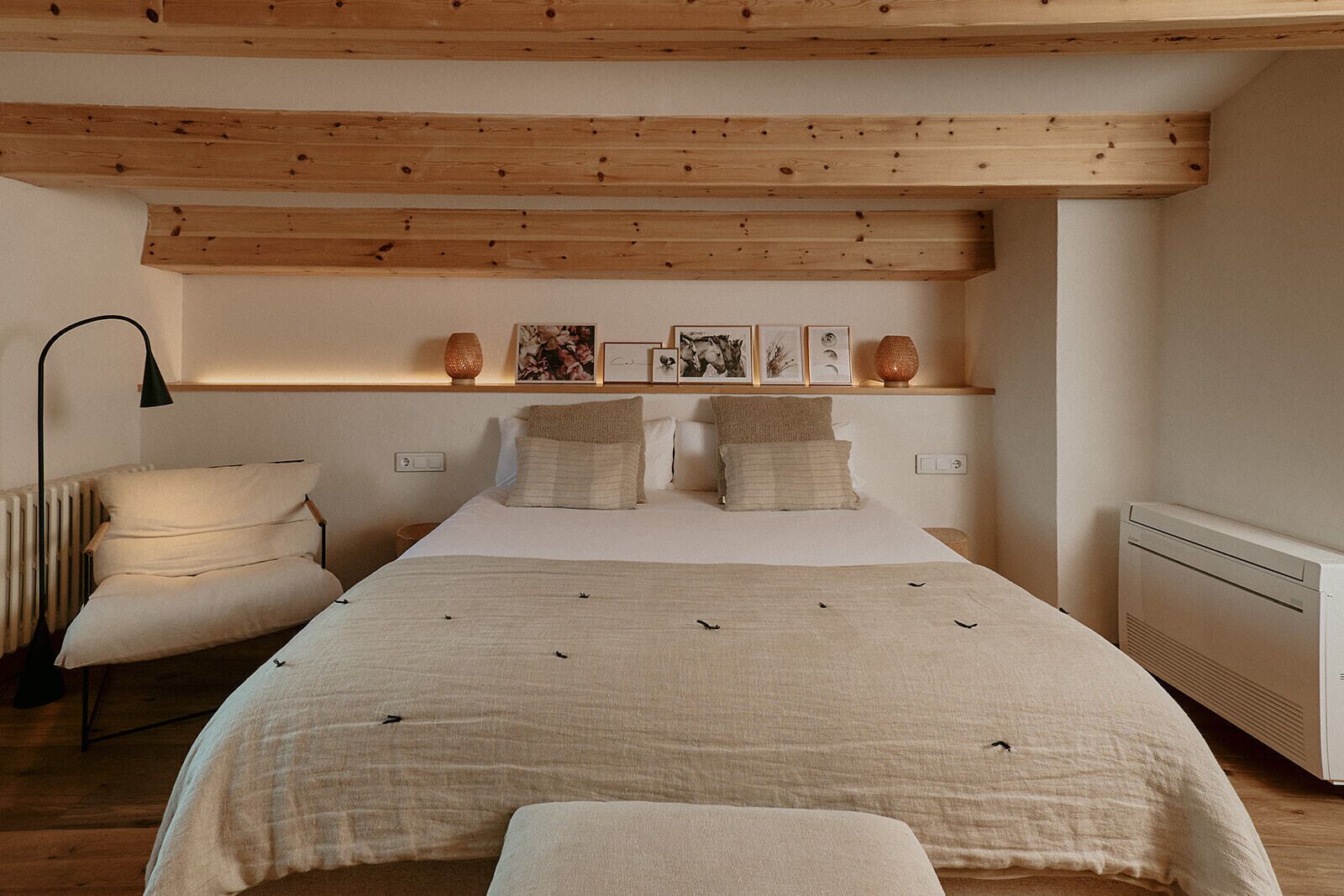 Un dormitorio con cama, almohadas, una silla, estante con fotografías y un radiador.