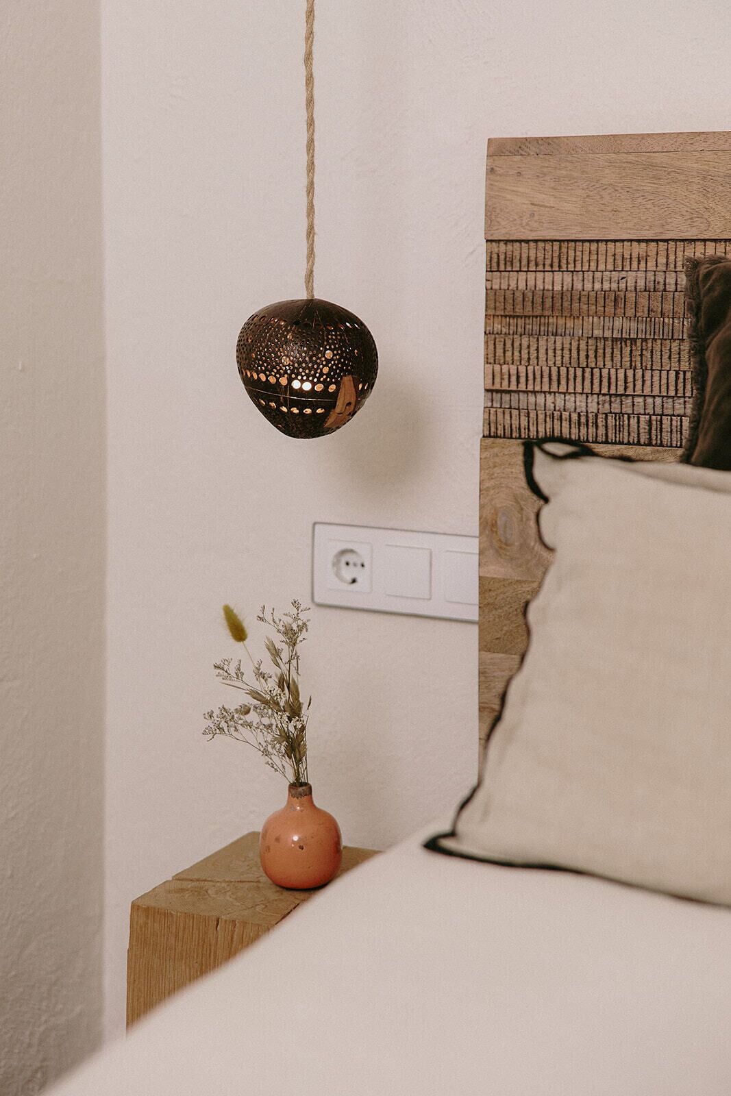 A bedroom with a wooden headboard, a hanging light fixture, a power outlet, and a small vase with dried flowers on a bedside table.