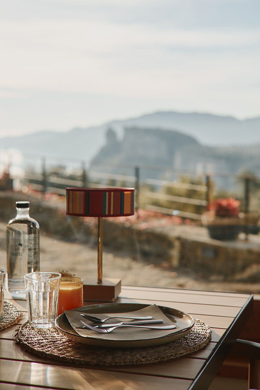 Una mesa dispuesta con un plato, cubiertos, una copa y una botella, con una lámpara y vistas a montañas al fondo.