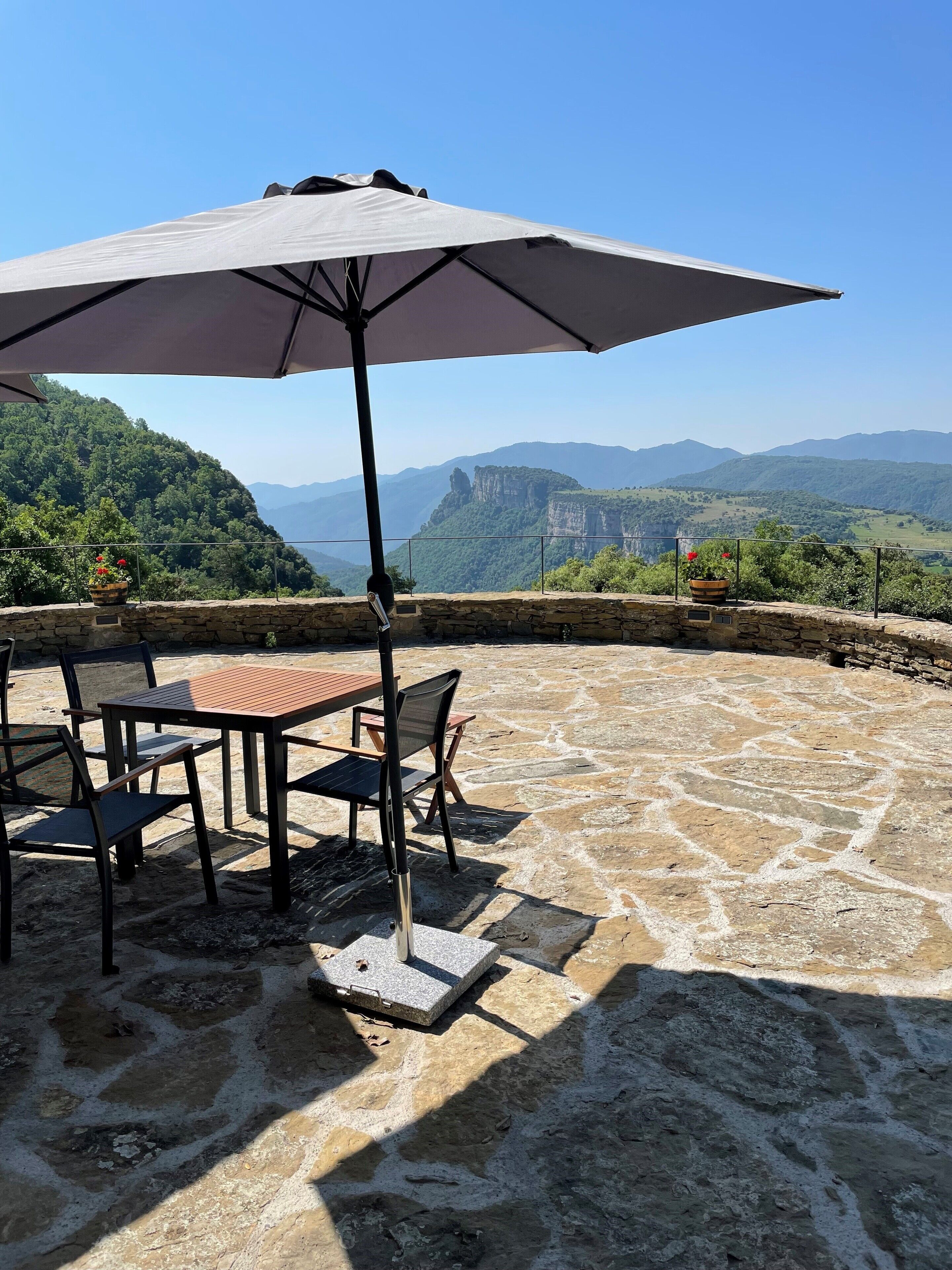 A patio with stone flooring and a table with chairs under a large umbrella, overlooking a mountainous landscape.