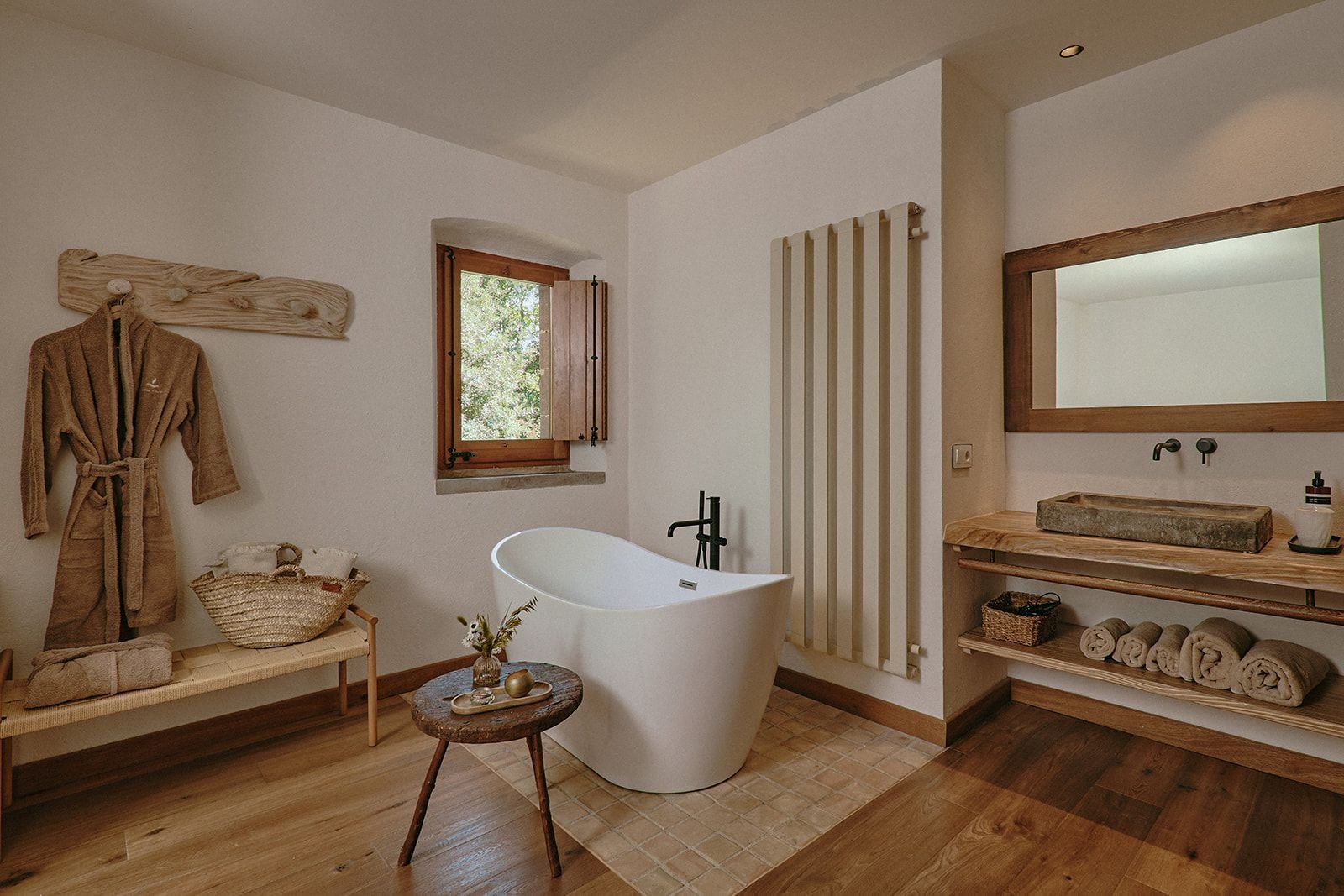 Un baño con una bañera independiente, repisas de madera con toallas, un banco de madera con una cesta y un ventanal con contraventanas.