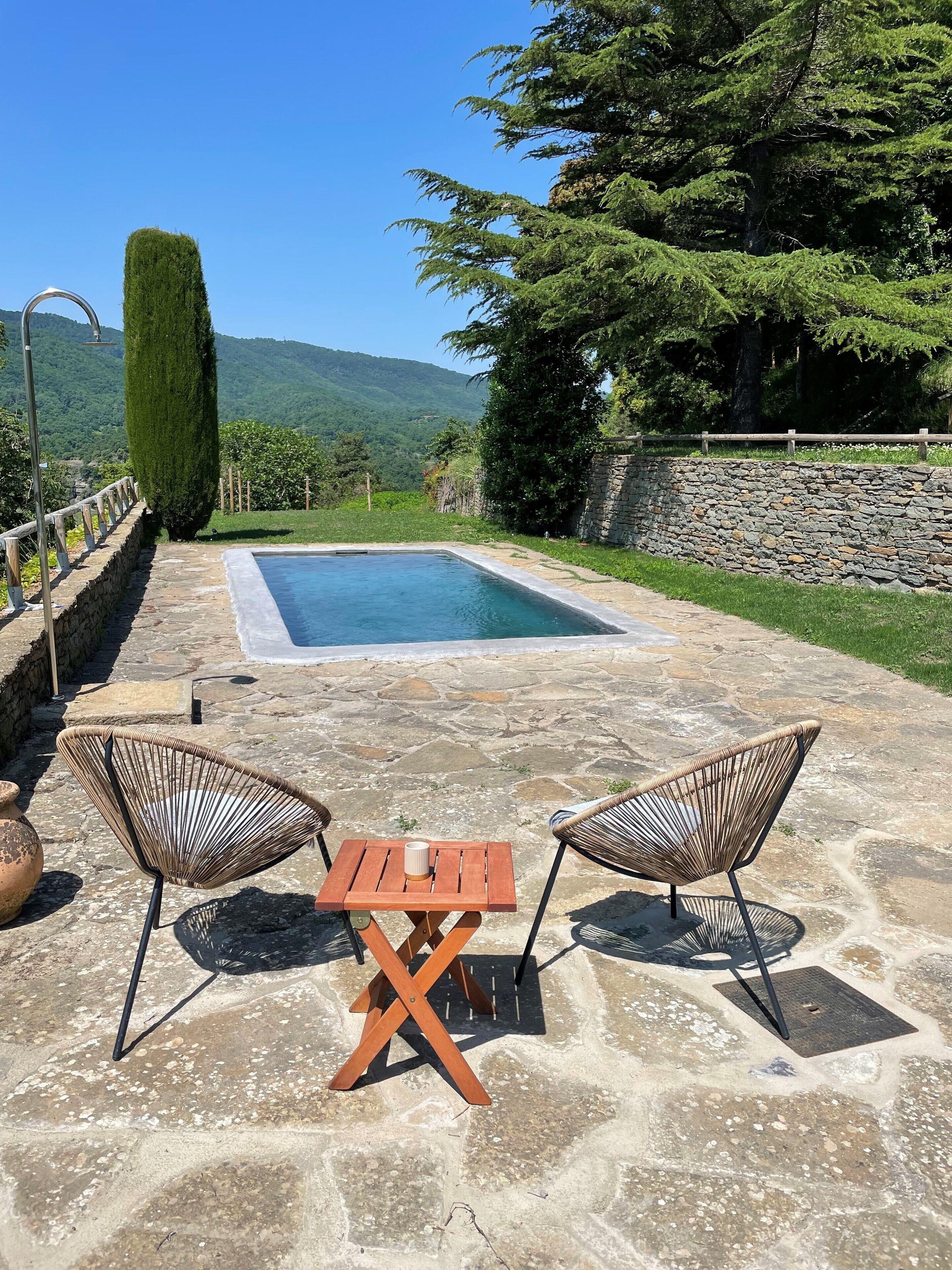 Un patio de piedra con una piscina, dos sillones de mimbre y una mesa de madera.