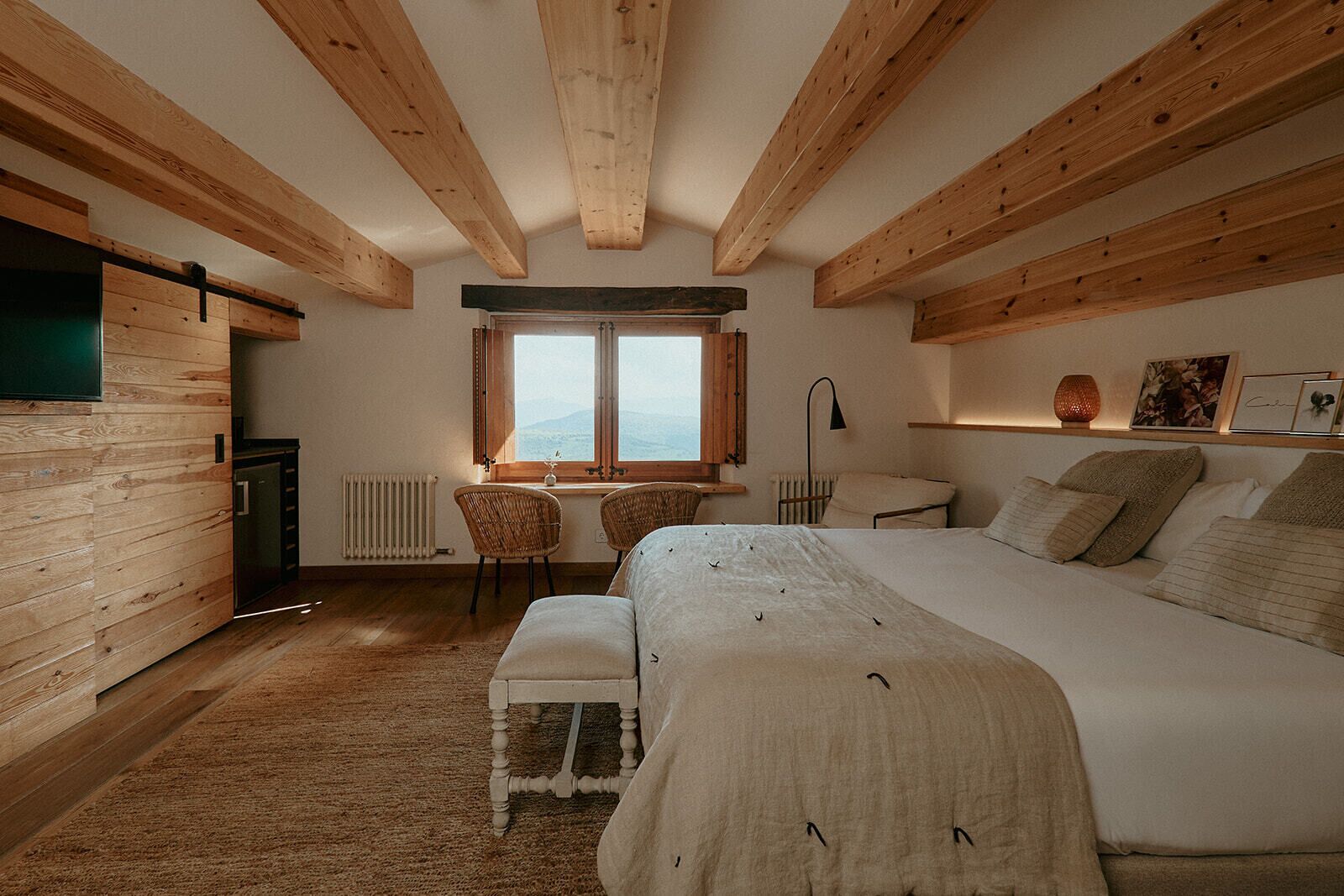 Een slaapkamer met een groot bed, houten balken, een raam, een bureau en een stoel.