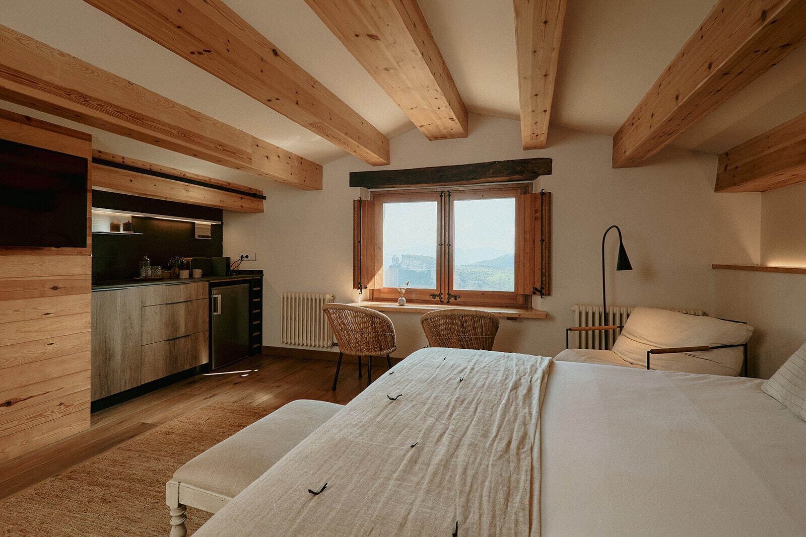 A bedroom with a bed, a window with a view, a desk with a chair, and a kitchenette.