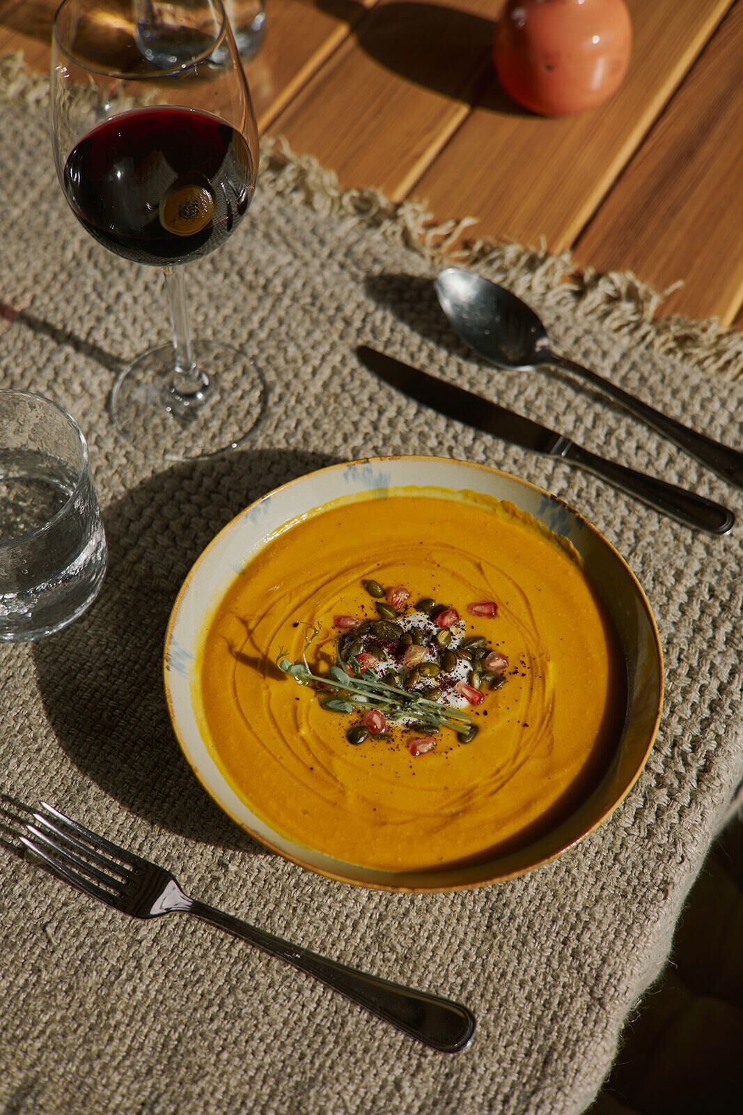 Une table avec un bol de soupe garni de graines et d’herbes, un verre de vin rouge et des couverts.