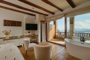 Een kamer met een bed, een bureau, een televisie, een groot raam en een badkamer met een bad.