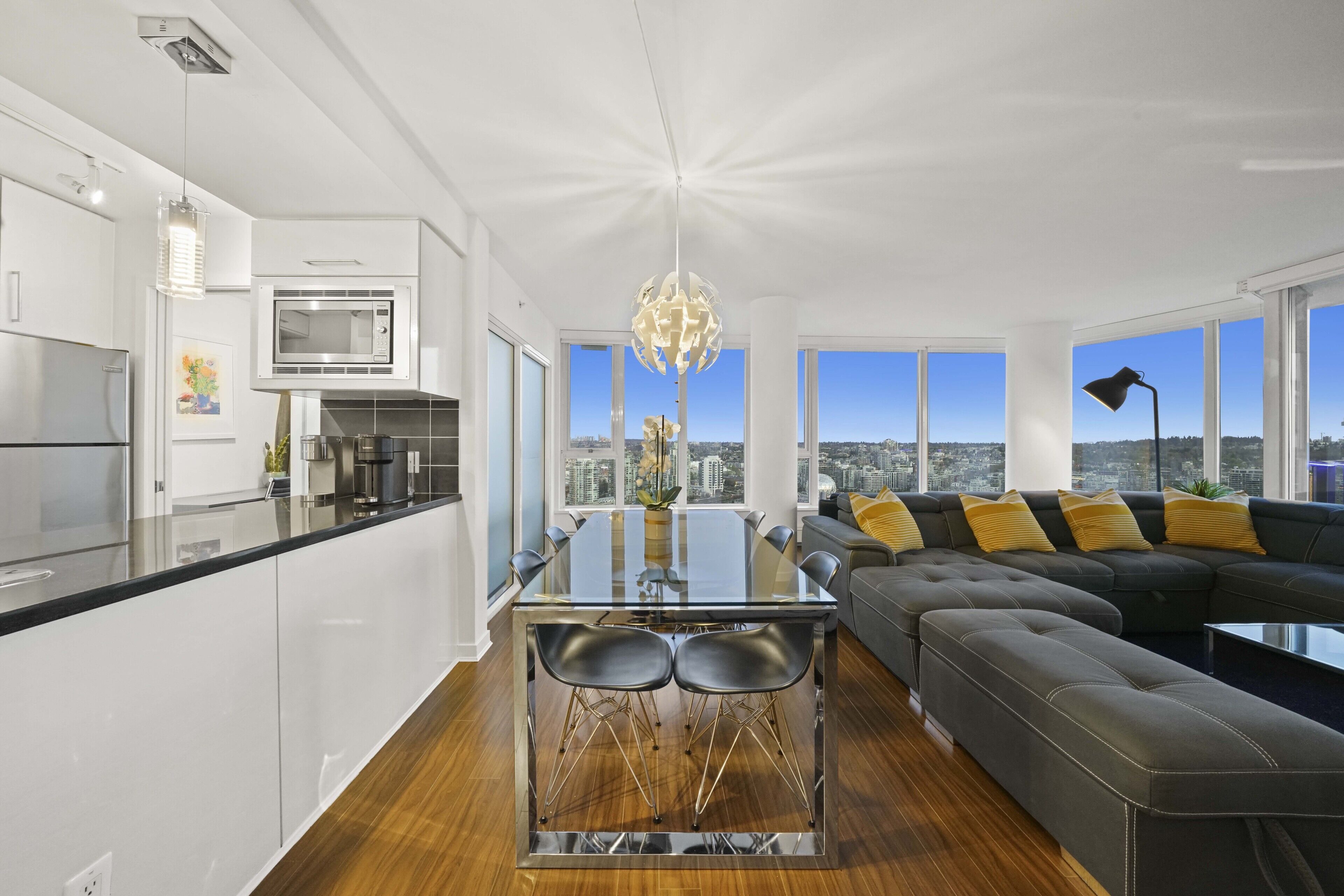 A modern kitchen with a glass dining table, a black sofa with yellow cushions, and a city view through large windows.