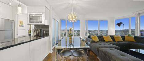 A modern kitchen with a glass dining table, a black sofa with yellow cushions, and a city view through large windows.
