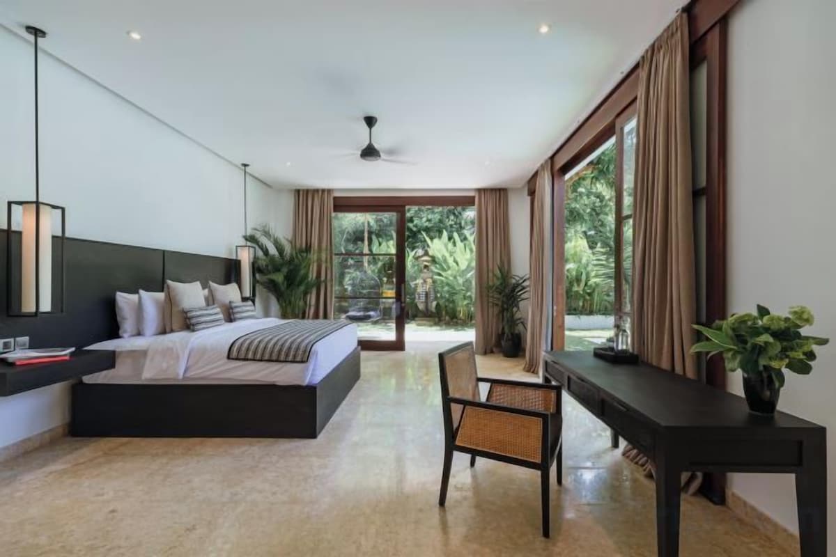 A modern bedroom with a large bed, a desk, and a chair. There is a view of greenery outside the window.
