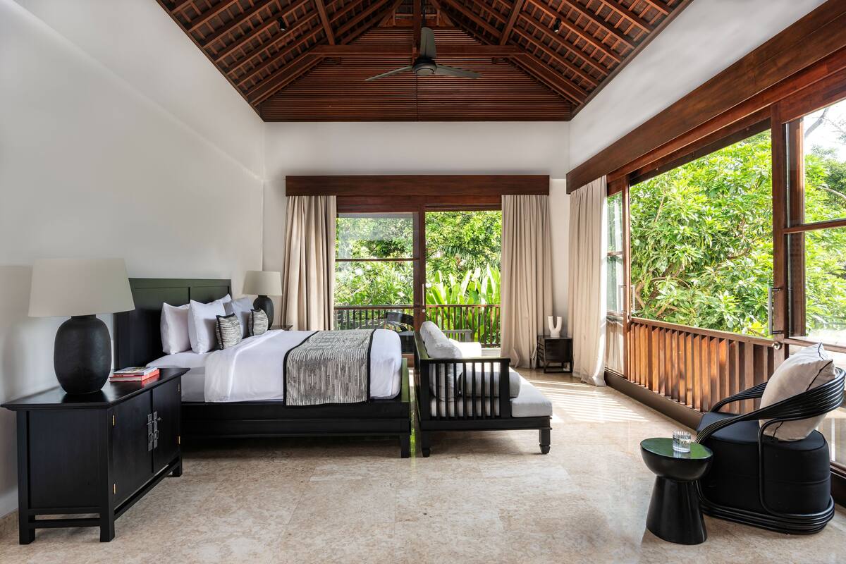 Un dormitorio con cama, mesita de noche, silla y ventanas amplias.