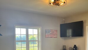 Uma sala de estar com piso de madeira, uma mesa redonda com um vaso de flores, uma poltrona amarela, uma TV de tela plana na parede e uma janela com vista para o verde.