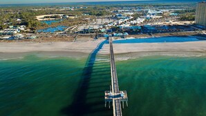 Luchtbeeld van een kustgebied met een lange pier die in het water reikt, omringd door gebouwen en een heldere lucht.