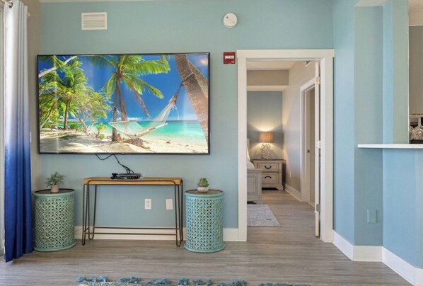 A room with a large TV on the wall, a wooden console table, two decorative planters, and a view into another room through an open door.