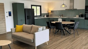 Una cocina moderna con armarios verde oscuro, una zona de comedor con una mesa redonda y sillas negras, y un sofá color beige con un cojín amarillo.