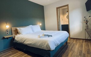 Una cama bien tendida con sábanas blancas y una toalla doblada, un cabecero de madera y una lámpara de pared.