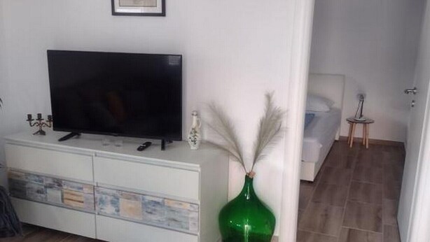 A living room with a flat-screen TV on a white cabinet, a green vase, and a door leading to another room.