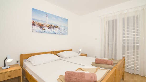 A bedroom with a wooden bed, white linens, a nightstand with a lamp, a painting of a lighthouse, and a window with blinds. - Apartment with sea view 1 (Pirovac)