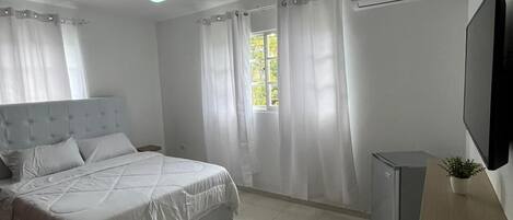 A bedroom with a bed, a ceiling fan, a window with curtains, a TV, a small refrigerator, and a plant.