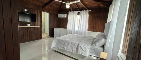 Un dormitorio amplio con una cama grande, vigas de madera en el techo, un ventilador de techo y un televisor montado en la pared.