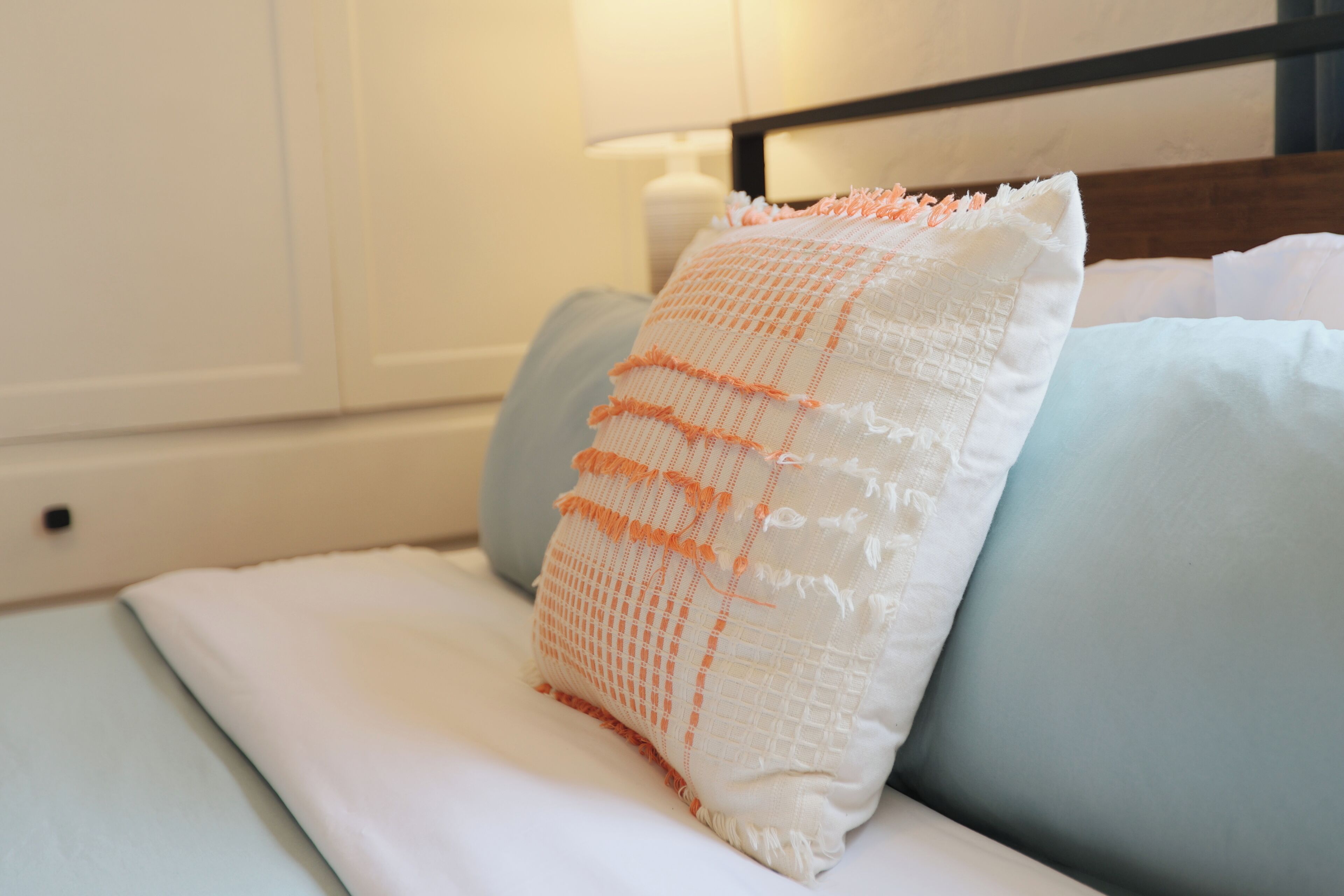 Un lit bien fait, avec un oreiller blanc et orange, un oreiller bleu clair et une tête de lit blanche.