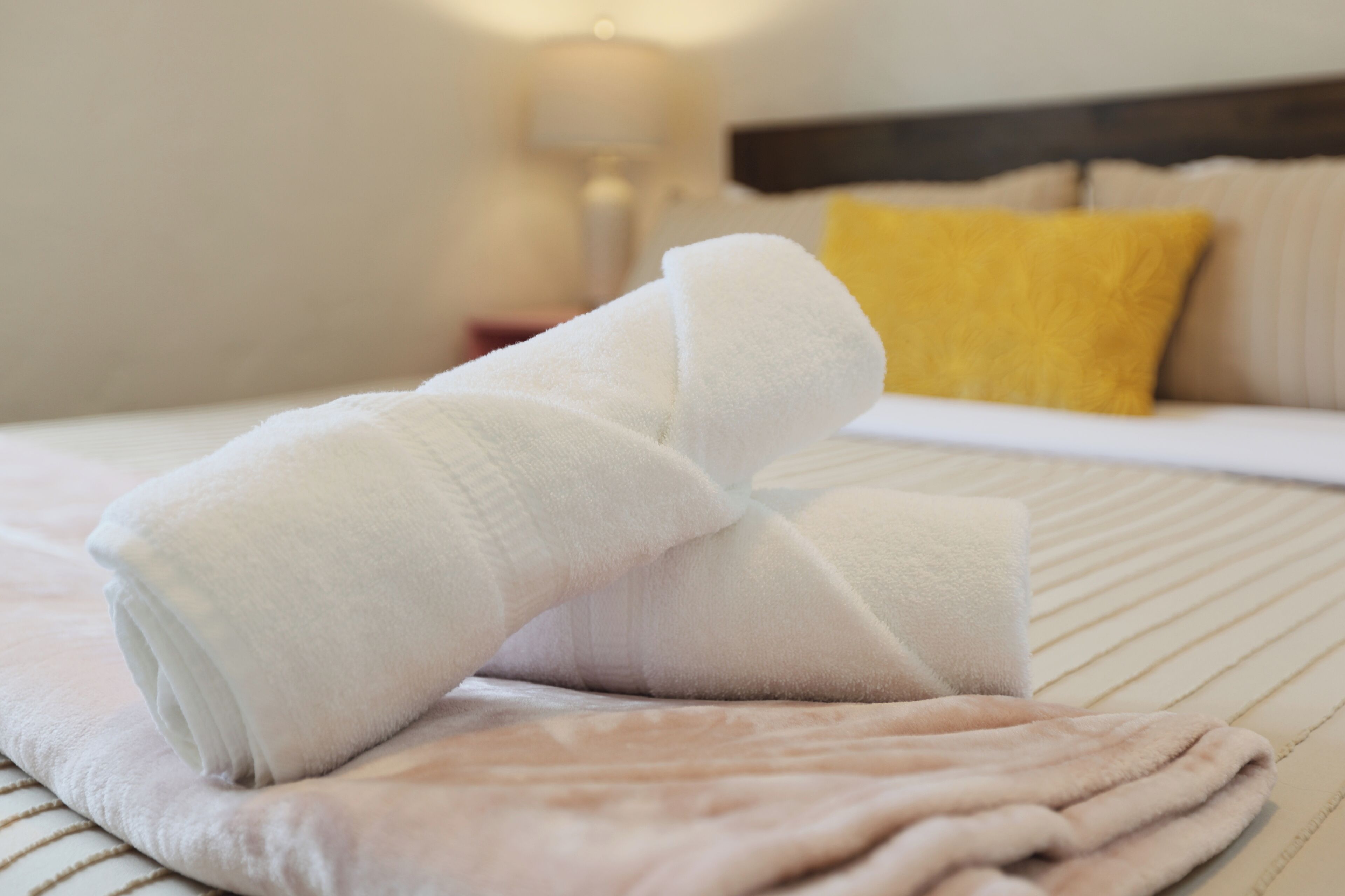 A neatly made bed with white and yellow pillows, a rolled white towel, and a beige blanket.