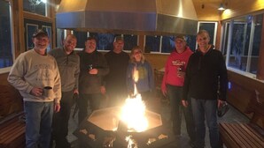 Un grupo de personas reunido alrededor de un brasero hexagonal en una habitación con paredes de madera y ventanas amplias.