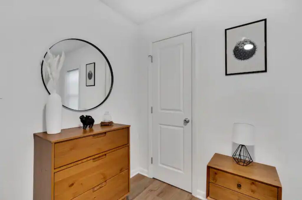 Una habitación con un aparador de madera, un espejo redondo, una puerta blanca y una mesita de noche con una lámpara.