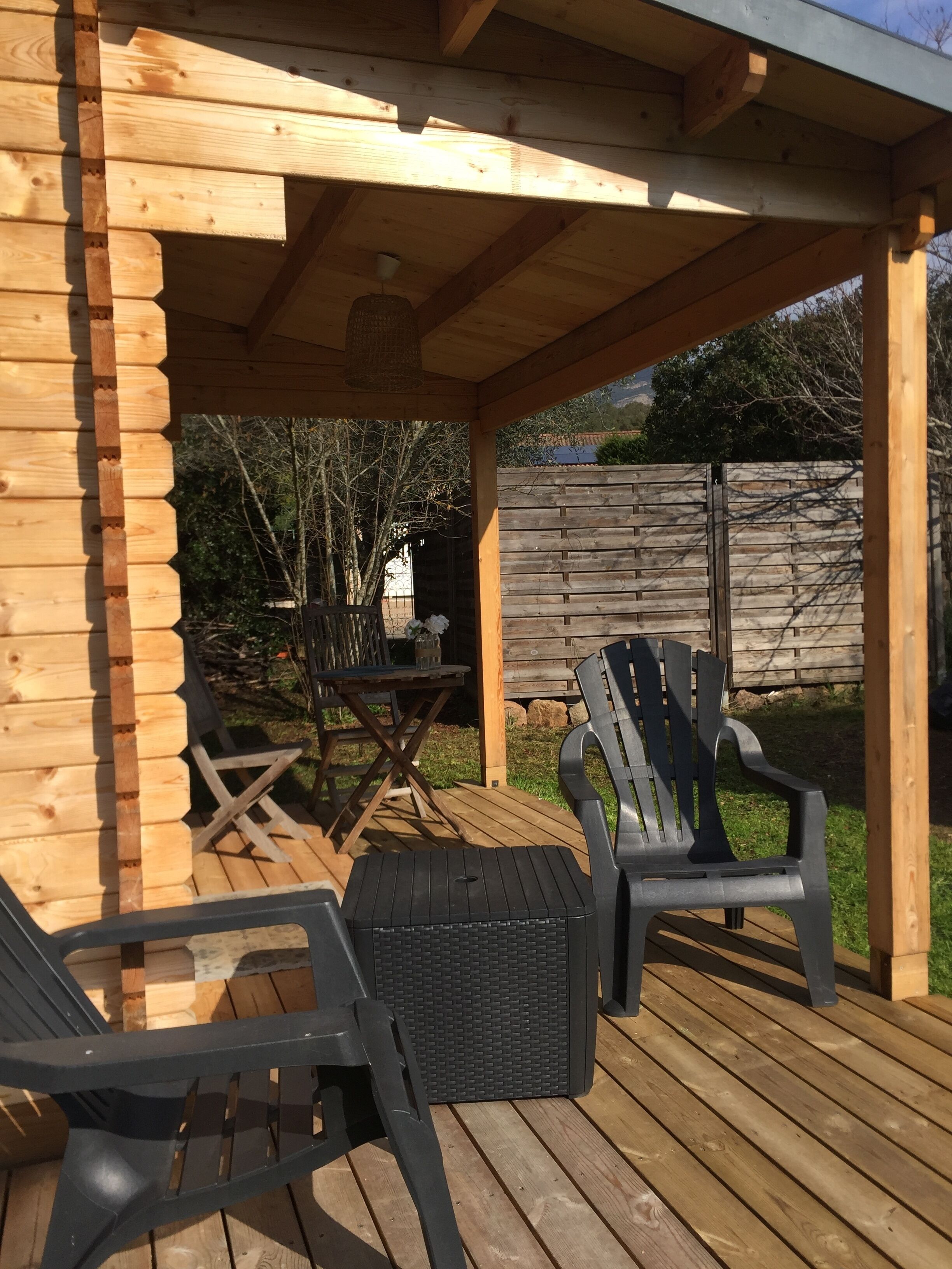 A wooden deck with two black chairs and a table, a pergola overhead, and a wooden fence in the background.