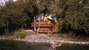 A cabin with a wooden deck and a small staircase leading to a second level, surrounded by trees and a rocky riverbank. - Charming Cottage on the River (South Bruce Peninsula)