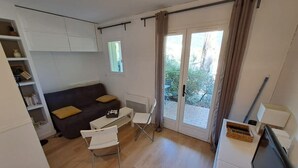 Ein kompakter Wohnbereich mit einer Couch, einem kleinen Tisch, einem Stuhl, einem Fernseher und einer Glastür, die zu einem Außenbereich führt.
