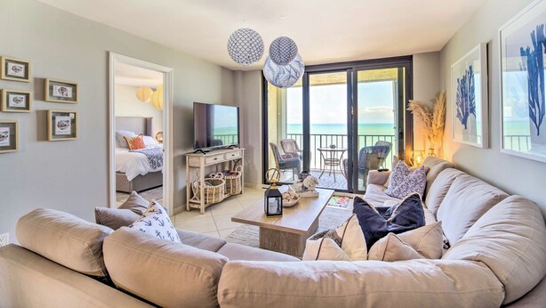A spacious living room with a large beige sofa, a coffee table, and a TV. There is a balcony with a view of the sea. - Lovely Fort Pierce Condo, Walk to Beach! (Fort Pierce)