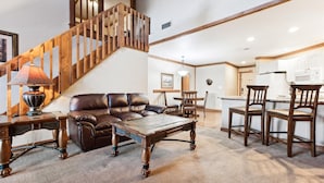 A spacious living area with a wooden staircase, leather sofa, wooden coffee table, and a dining area with wooden chairs and a kitchen in the background.