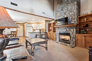 A spacious living room with a stone fireplace, wooden shelves, and a kitchen in the background.