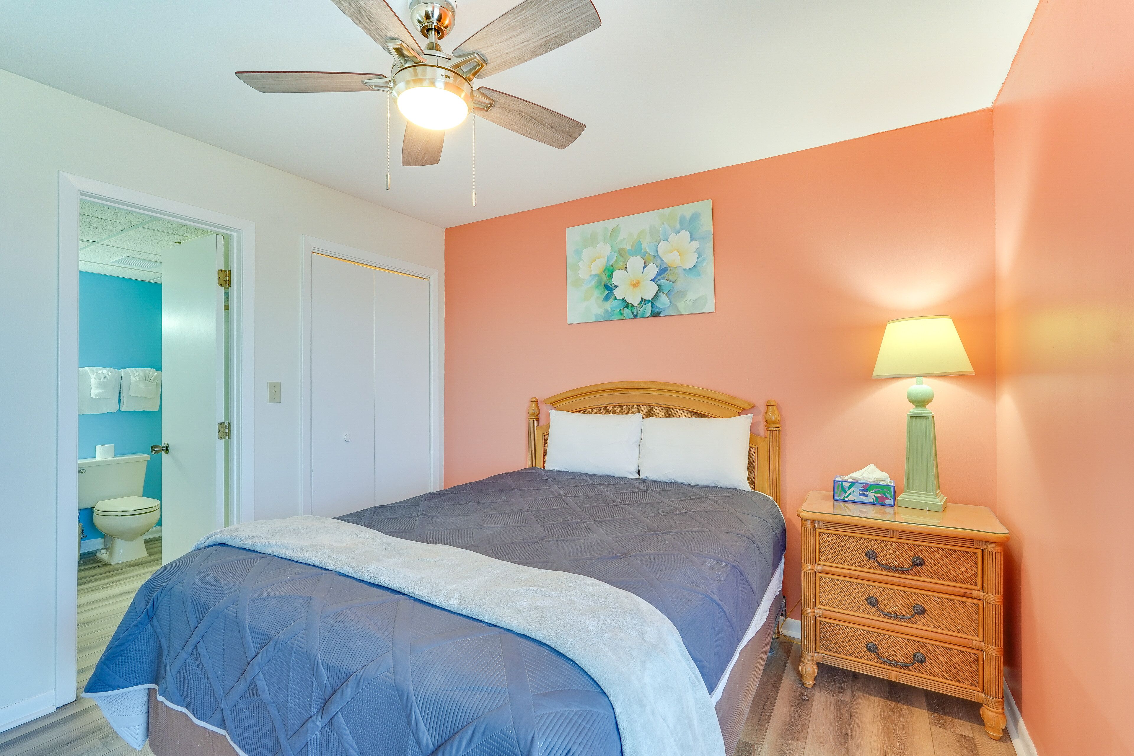 A bedroom with a bed, a nightstand with a lamp, a ceiling fan, and a door leading to a bathroom.