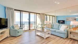 A modern living room with a large flat-screen TV, a light blue sofa, a white coffee table, and a balcony with a view of the sea. - PCB Vacation Rental w/ Sweeping Ocean Views! (Panama City Beach)