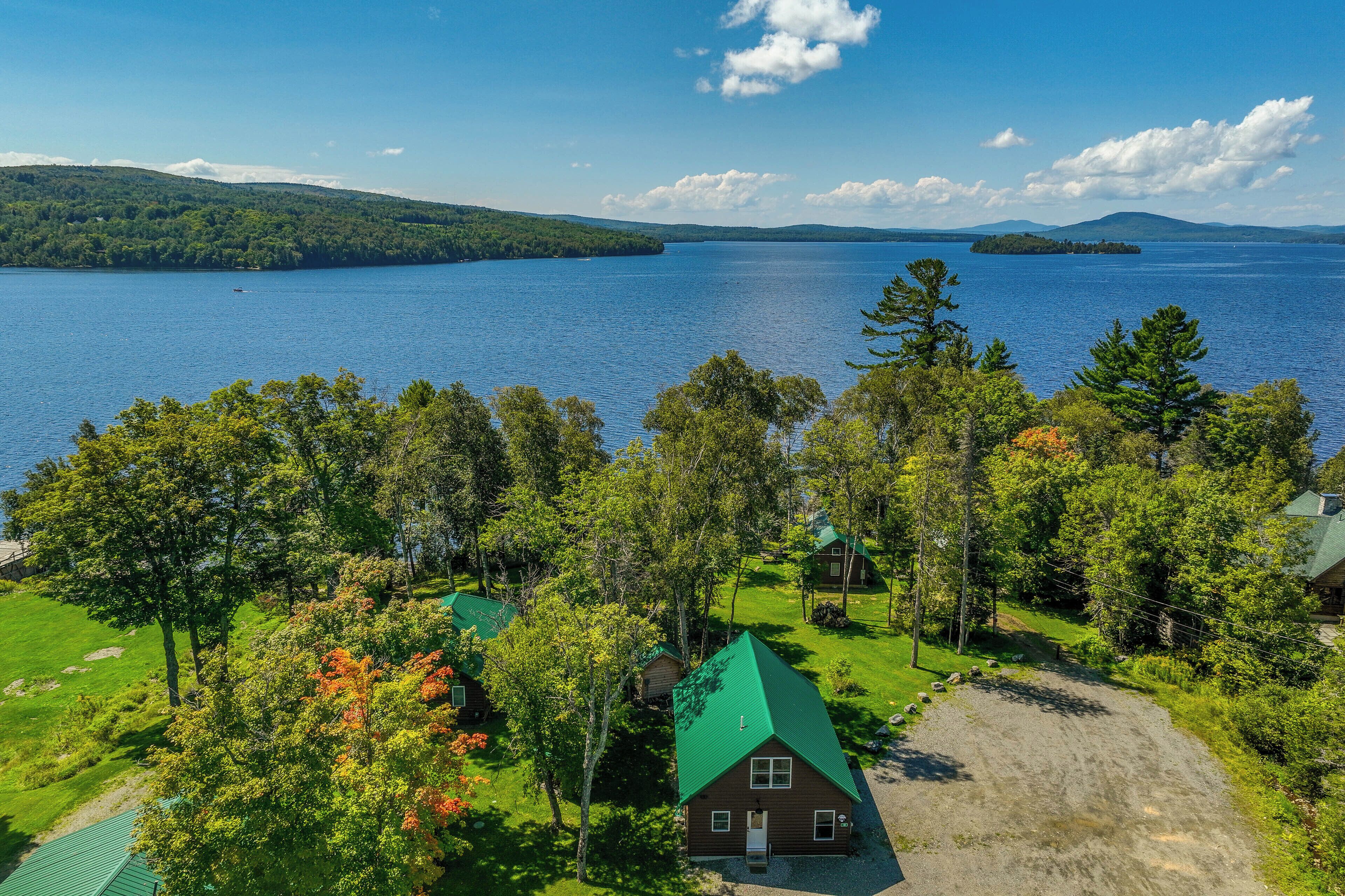 A cabin with a green roof surrounded by trees, overlooking a large body of water and distant mountains.
